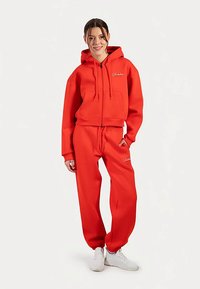 Sweat à capuche rouge avec fermeture éclair à l'avant, cordon de serrage et poches avant, assorti à un pantalon de jogging assorti. Les deux ont une texture de tissu lisse.