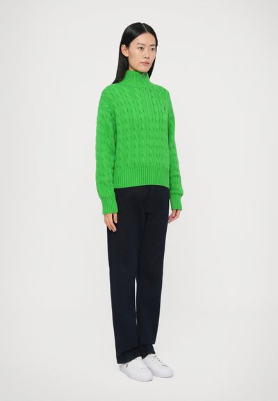 Zelený sveter s károvaným vzorom, vysokým golierom, rebrovanými manžetami a lemom; v kombinácii s tmavomodrými nohavicami a bielymi teniskami.