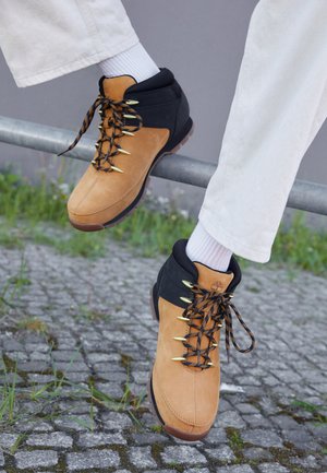 Brown and black hiking boots with suede upper, gold eyelets, and patterned laces, displayed on a railing over cobblestones. White socks visible.