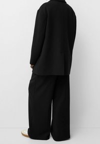 Person som bär en överdimensionerad svart jacka och vida svarta byxor, stående mot en enkel ljus bakgrund med vita sneakers synliga.