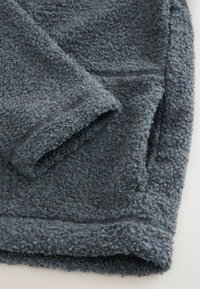 Rukáv a kapsa tmavě šedé fleecové bundy s texturou a kudrnatou vlněnou látkou na světlém pozadí.