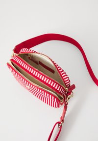 Czerwono-biała torba crossbody w paski, z dwoma zamykanymi na zamek kieszeniami, złotymi akcentami i czerwoną tkanym paskiem. Wnętrze zawiera kieszenie na karty.