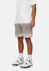 Shorts en mélange de coton gris avec un effet dégradé, texte brodé en bleu foncé, associés à des baskets blanches et grises et à un T-shirt blanc ample.