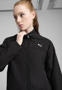 Črna lahka jakna z visokim ovratnikom, zadrgo in srebrnim logotipom Puma na prsih. Gladka tkanina z atletskim dizajnom.