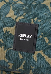 Tissu à motif floral en vert et beige avec une finition texturée. Présente un carré noir étiqueté "REPLAY SINCE 1981."