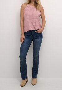 Donna con un top rosa senza maniche a pizzo, jeans di denim blu, stivali alla caviglia in suede beige e un bracciale dorato, in piedi contro un muro semplice.