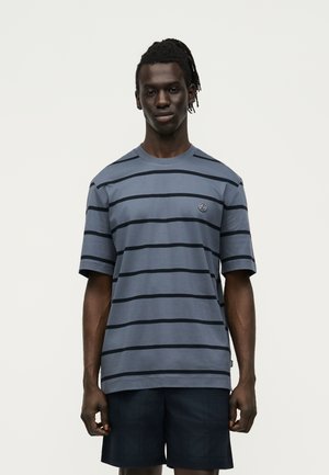 Homme à la peau foncée et aux dreadlocks portant une chemise bleu-gris à manches courtes avec des rayures horizontales noires et un short foncé, debout sur un fond clair.