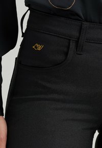 Jeans noirs taille haute en denim, présentant une texture subtile, avec un logo "LW" brodé en or sur la poche avant.