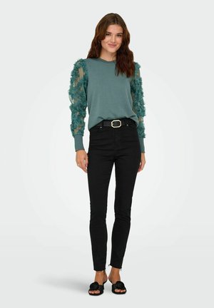 Sweatshirt teal avec des manches transparentes texturées florales, associé à un jean skinny noir et des sandales noires. Design simple, coupe décontractée.