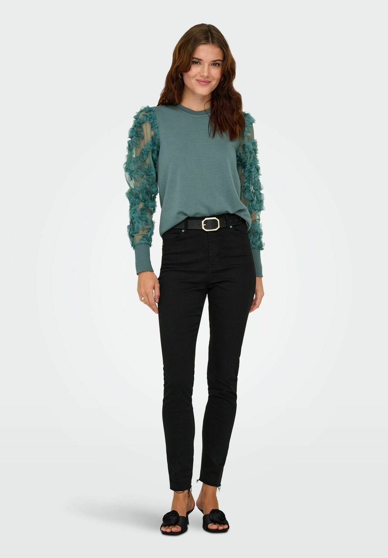 Felpa turchese con maniche trasparenti a motivo floreale, abbinata a jeans skinny neri e sandali neri. Design semplice e vestibilità casual.
