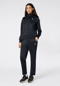 Zwart atletisch trainingspak van gladde stof, met een jas met rits en opstaande kraag en een broek met zijzakken, zichtbare logo-accenten.