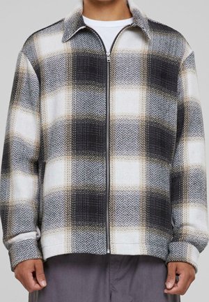 Homme portant une veste zippée à carreaux noirs, blancs et beiges, avec manches longues et col, par-dessus une chemise blanche.