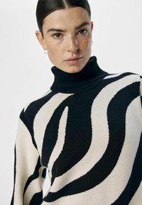 Pull noir et crème à col roulé avec un motif en ondulations. Texture en tricot douce avec des accents côtelés au niveau du col et des manches longues.