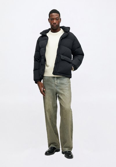 Juodas pūkinis švarkas su grietinėlės spalvos megztiniu, dėvimas su laisvais išblukusiais džinsais ir juodais bateliais, asmuo, stovintis su viena ranka kišenėje prieš baltą foną.