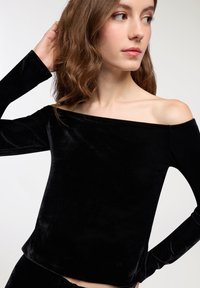 Top nero off-shoulder realizzato in morbido velluto, con maniche lunghe e una texture liscia. Design semplice e aderente senza decorazioni.