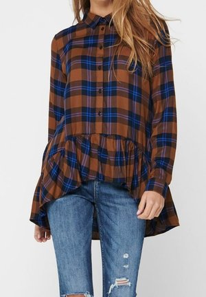 Femme portant une chemise à carreaux marron et bleue boutonnée avec un ourlet à volants, associée à un jean bleu déchiré.