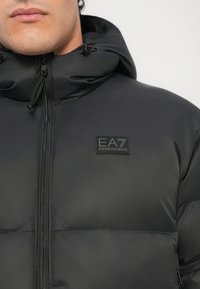 Mann trägt eine schwarze gepolsterte Jacke mit Kapuze und einem EA7 Emporio Armani Logo-Patch auf der Brust.
