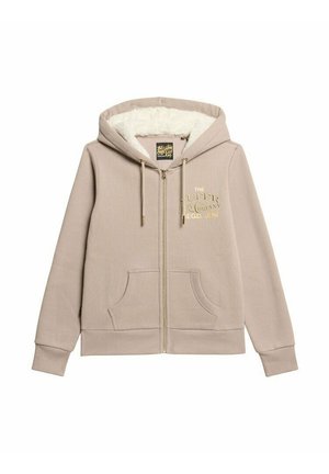 Beige Zip-Hoodie mit einem plüschigen weißen Innenfutter, großen Fronttaschen und einem goldenen gestickten Logo auf der Brust. Verfügt über Kordeln an der Kapuze.