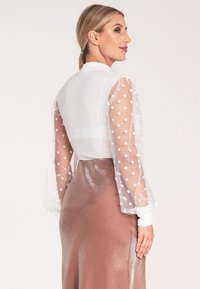 Przezroczysta biała bluzka z bufkami w kropki, zestawiona z błyszcząca brązową spódnicą. Posiada klasyczny kołnierzyk i mankiety na guziki.