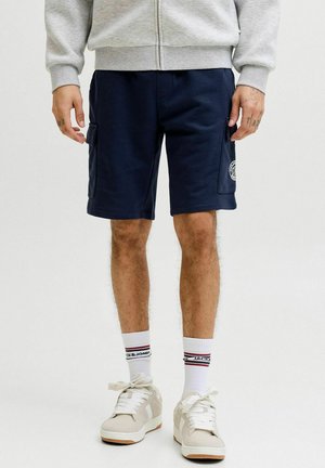 Mężczyzna ubrany w granatowe szorty cargo, białe skarpetki z logo, beżowe tenisówki oraz jasnoszarą bluzę z zamkiem, stoi z ramionami w luźnej pozycji.