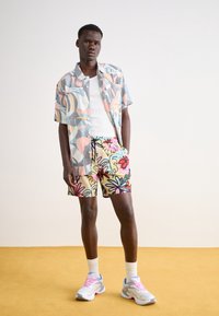 Krátký rukáv má košile s vícolorovým abstraktním vzorem; šortky jsou světlé s výraznými květinovými motivy; na nohou má bílé tenisky s růžovými akcenty.