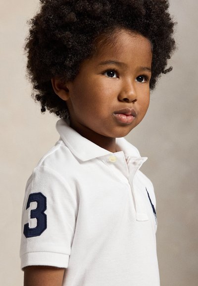 Jeune enfant aux cheveux bouclés portant un polo blanc avec un numéro trois bleu marine sur la manche, regardant légèrement vers la droite.
