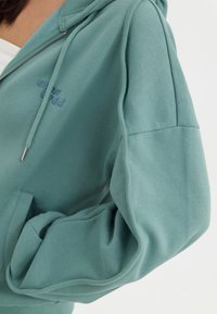 Persona che indossa una felpa con zip color turchese, con tasca frontale e lacci, mano casualmente nella tasca, testo ricamato vicino al petto.