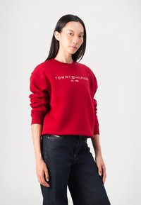 Červená mikina s ribbenými manžetami a výstřihem, s bílým textem „TOMMY HILFIGER EST. 1985“ na přední straně. Stylizováno s tmavými džíny.