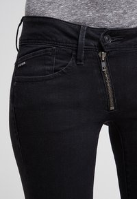 Black denim jeans with a slim fit, featuring a front zippered closure, subtle branding on the pocket, and a smooth texture.