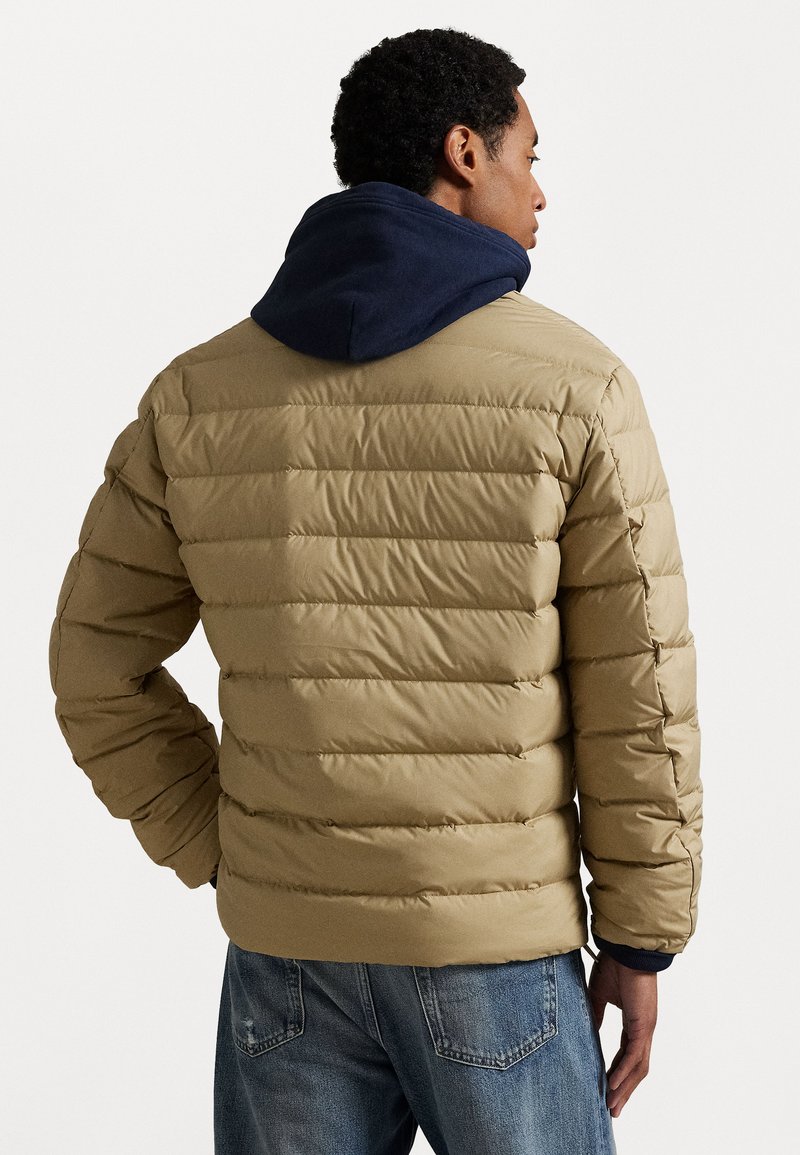 Beige gewatteerde jas met horizontale stiksels, voorzien van een marineblauwe capuchon. De jas heeft een relaxte pasvorm, elastische manchetten en een soepele textuur.