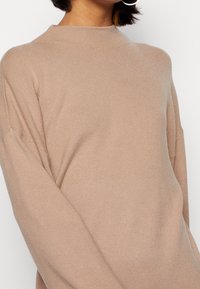 Pull en tricot beige avec un col montant, une coupe décontractée et des manches longues. Texture douce avec des accents côtelés au col et aux poignets.