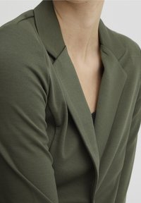 Olivově zelený blazer s výstřihem ve tvaru klop. Měkká, texturovaná látka zakrývá ramena a výstřih, čímž zdůrazňuje přiléhavý siluetu.