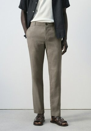 Mężczyzna w beżowych spodniach o prostym kroju, ciemnobrązowych skórzanych sandałach, białej koszulce i rozpiętej czarnej koszuli z krótkim rękawem, stojący z rękami w kieszeniach.