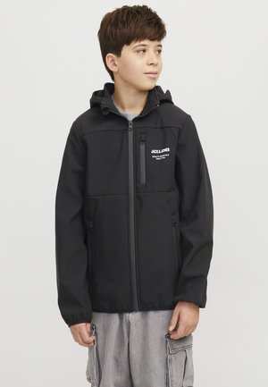 Chlapec oblečený v čiernej Jack & Jones mikine na zips s kapucňou a svetlosivých cargo nohaviciach, pozerá sa na stranu na jednofarebnom pozadí.