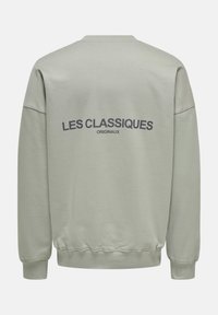 Hellgrüner Pullover mit geripptem Kragen und Bündchen. Auf der Rückseite sind "LES CLASSIQUES" und "ORIGINAUX" in dunkelgrau gedruckt. Weicher Stoff.