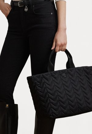 Svart quiltad tote-väska med chevronmönster, mjukt tyg, dubbla handtag och logodetaljer. Buren med svarta jeans och höga stövlar.