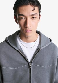 Mladý človek s krátkymi kučeravými vlasmi, nosiaci strieborný reťazový náhrdelník, malé krúžkové náušnice, šedú zip-up mikinu a biele tričko pred bielym pozadím.