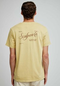 Lys gul, casual t-shirt med korte ærmer, med "Scalpers Co." trykt i mørkebrun på ryggen, teksturen er blød og glat.