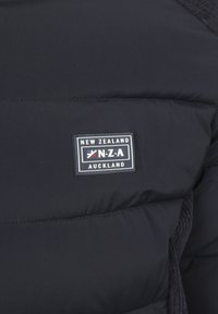 Zwarte gewatteerde jas met een quilted textuur, voorzien van een rechthoekig logo-opdruk dat "NEW ZEALAND AUCKLAND" en "N.Z.A." in wit en rood zegt.