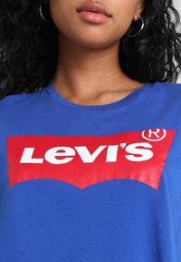 Blå bomull t-shirt med en stor röd Levi's-logga, med vit text och ett distinkt krökt designelement under loggan.