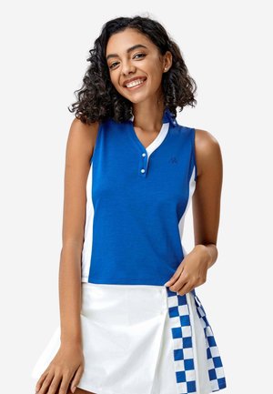 Femme souriante aux cheveux bouclés portant un top bleu sans manches et une jupe blanche avec des panneaux latéraux à carreaux bleus.