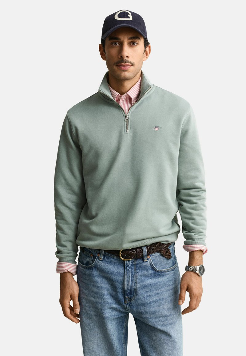 Homme portant une casquette bleu marine avec un "G", un sweat à col zippé vert sauge sur une chemise rose à rayures, un jean bleu, une ceinture tressée marron et une montre argentée.