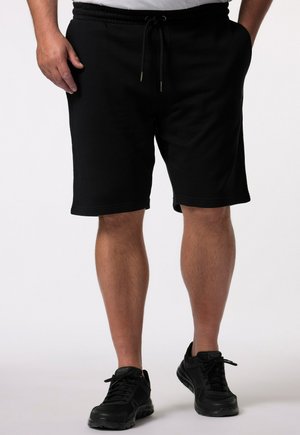 Person trägt schwarze Shorts mit Kordelzug und schwarze Sneakers und steht vor einem schlichten weißen Hintergrund.