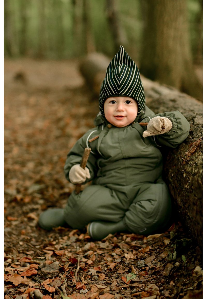 Zeleni podložen snežni kombinezon z elastičnimi manšetami, skupaj s črno-belim črtastim klobukom. Otrok drži palico in sedi na hlodu v gozdu.