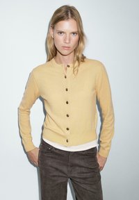 Gul stickad kavaj med rund halsringning, sex bruna knappar och en figursydd design. Bärs över en vit skjorta och kombineras med mörka jeans.