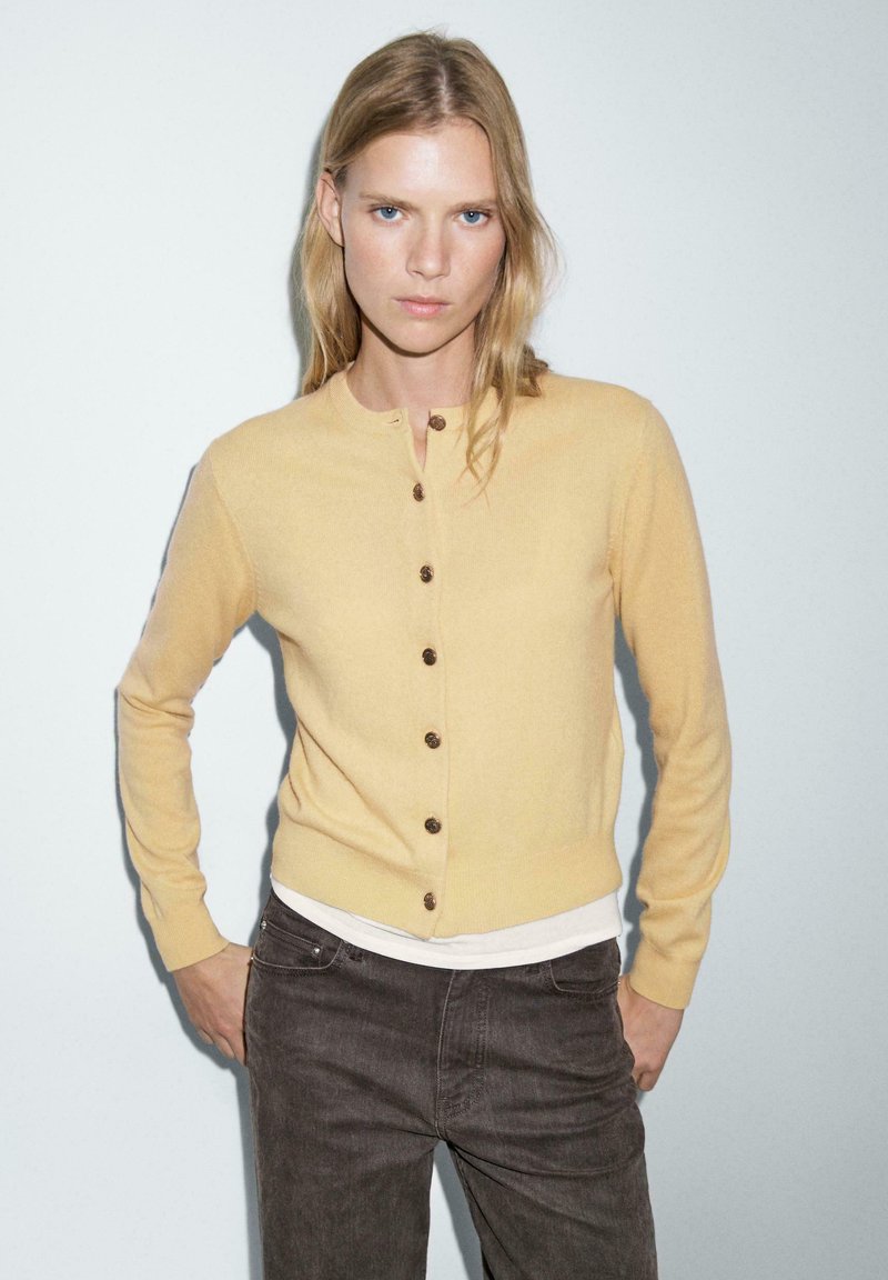Gul stickad kavaj med rund halsringning, sex bruna knappar och en figursydd design. Bärs över en vit skjorta och kombineras med mörka jeans.