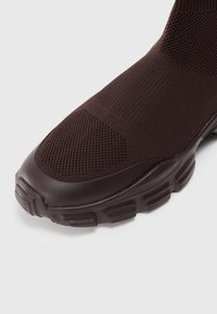 Hnědá sportovní obuv s pleteným svrškem a texturovanou síťovinou, elegantní gumovou podrážkou a zaobleným designem špičky pro flexibilitu.