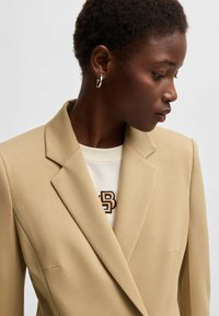Beige bleiser yang disesuaikan terbuat dari kain halus, dilengkapi dengan bahu terstruktur, kerah terpotong, dan satu saku dada.