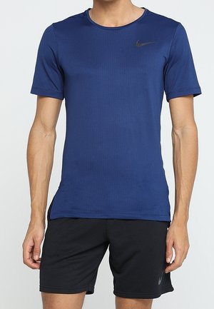 Man draagt een blauw Nike T-shirt met korte mouwen en zwarte Nike sportshorts, staat met ontspannen armen langs het lichaam.