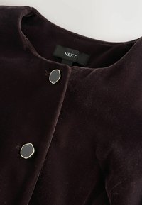 Tume lilla sametist jope ümmarguse kaelusega, millel on kaks ovaalset musta nuppu kuldse raami sees, millel on silt "NEXT". Sile tekstuur.